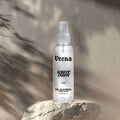 Veena White Oudh alcohol-free attar spray 100ml bottle on a rugged stone podium with soft leaf shadow overlays.