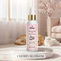 Veena Cherry Blossom Pet Perfume for Dogs and Cats 100ml spray bottle resting on a white marble platform with blooming pink cherry blossom trees and a white cat in the background.