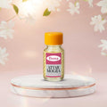 Veena Premium Attar Mogra 2ml miniature glass bottle standing on clean white marble podium with soft pink jasmine graphics backdrop