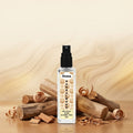 Veena Sandalwood Ultra Luxury Attar 8ml alcohol-free spray bottle centered amongst raw sandalwood logs and wood shavings on a warm wooden background.