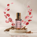 Veena Attar Ruh Gulab premium concentrated pure rose perfume oil bottle with a gold cap, set on a polished white marble pedestal surrounded by swirling pink rose petals, natural wood chips, and elegant curls of aromatic smoke.