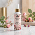 Veena Rose Petals Potpourri Spray 100ml bottle displayed on a polished white marble surface surrounded by pink roses and delicate buds – Soft Floral Elegance Home Fragrance.