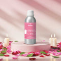 Veena Rose Fragrance for Candle Making aluminum bottle sitting on a pink round podium surrounded by burning candles and loose rose petals.